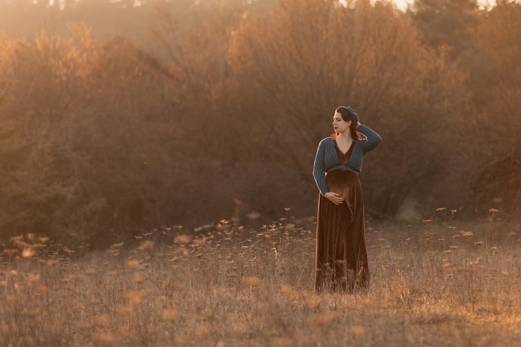 Schwangere Frau mit roten Haaren im braunen Samtkleid und petrolfarbener Weste im Abendlicht auf einer Wiese, stimmungsvolle Outdoor-Fotografie in Ludwigsburg