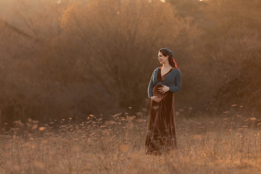 Schwangere Frau mit roten Haaren im braunen Samtkleid und petrolfarbener Weste im Abendlicht auf einer Wiese, stimmungsvolle Outdoor-Fotografie in Stuttgart Winterlocatin in weichen abendlicht