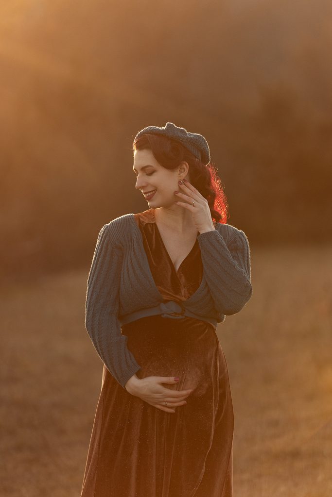 Schwangere Frau mit roten Haaren im braunen Samtkleid und petrolfarbener Weste im Abendlicht auf einer Wiese, stimmungsvolle Outdoor-Fotografie in Stuttgart