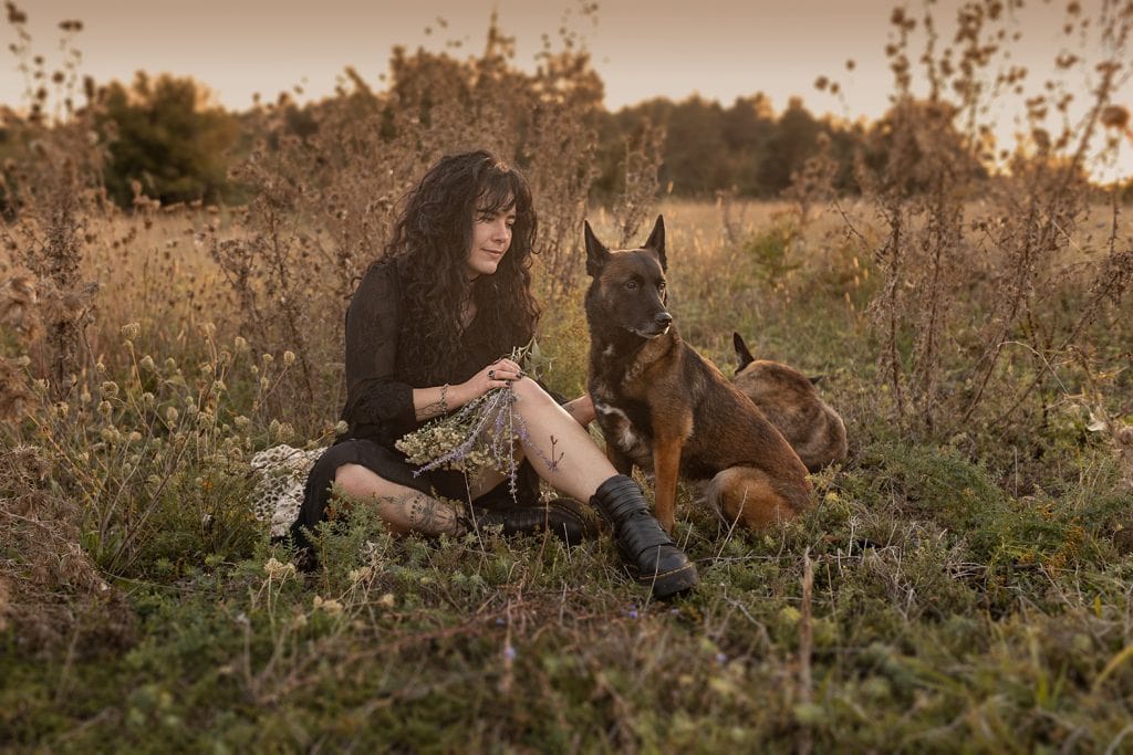 Frau mit langen schwarzen Locken sitzt mit ihrer Malinois-Hündin auf einer herbstlichen Wiese bei einem Outdoor-Fotoshooting in Stuttgart.