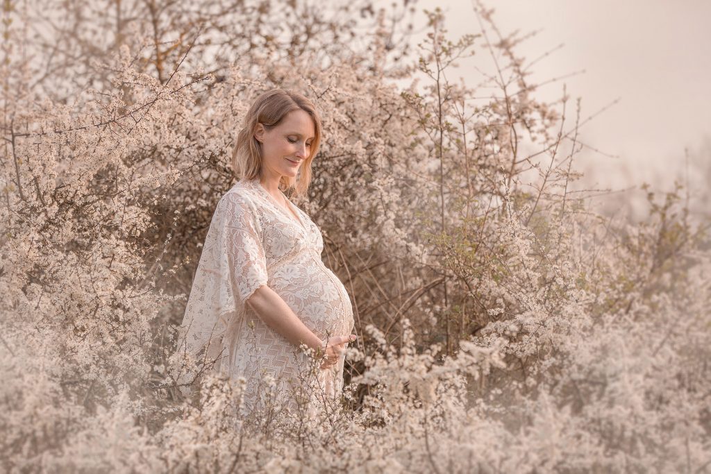 Schwangere Frau in weißem Spitzenkleid steht inmitten weißer Schlehenblüten, natürliche Schwangerschaftsfotografie in Stuttgart, liebevolle Outdoor-Aufnahme
