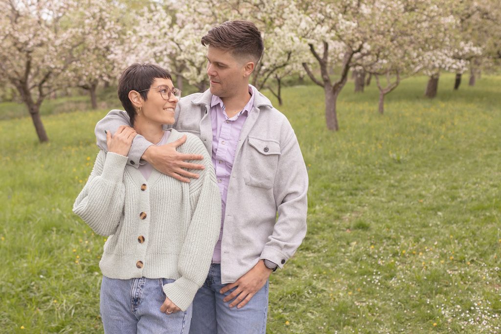 Paar steht auf blühender Obstbaumwiese, er legt Arm von hinten um sie, beide schauen sich liebevoll an, Hunde im Hintergrund – Paarshooting mit Hund Stuttgart.