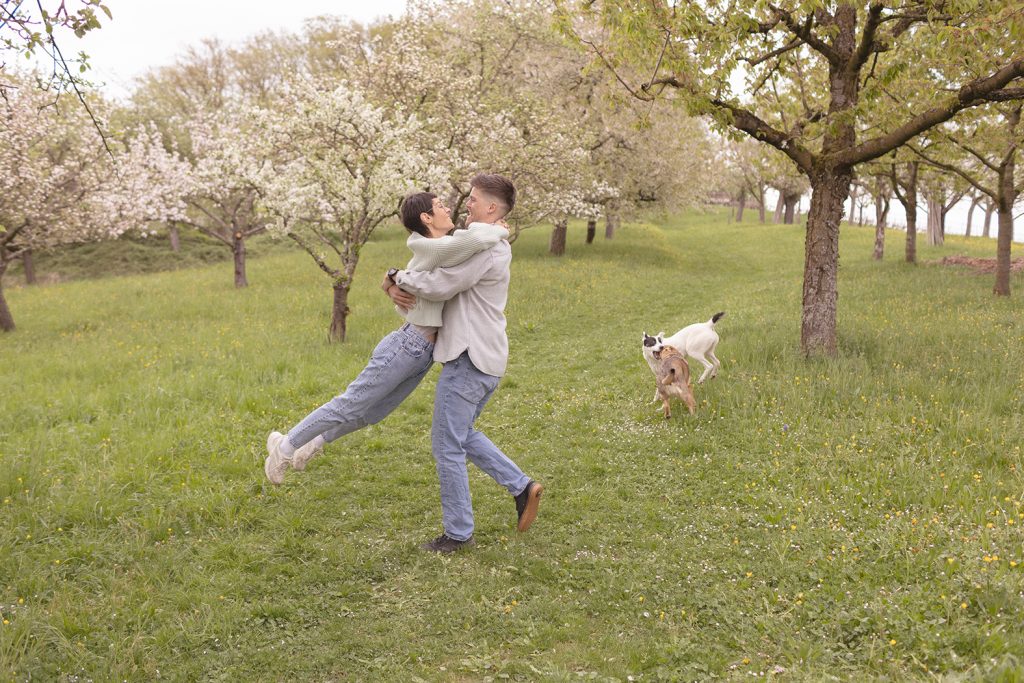 Mann dreht Frau im Kreis auf grüner Wiese, Hunde spielen im Hintergrund auf Obstbaumwiese – Paarshooting mit Hund Stuttgart.