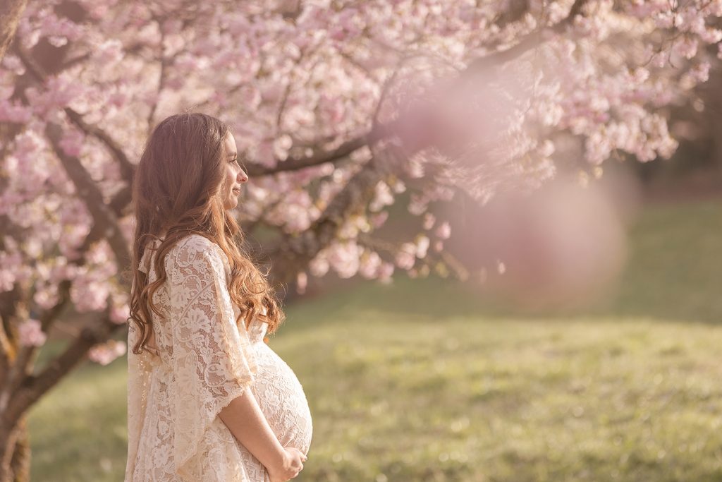 Schwangere Frau in hellem Spitzen Babybauchkleid blickt verträumt in die Ferne vor rosa Kirschblüten – elegantes Frühlings-Babybauch-Fotoshooting Stuttgart.