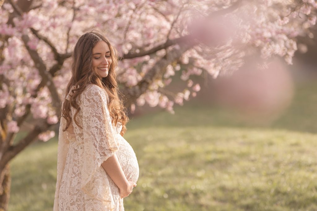 Frau in Spitzen-Babybauchkleid zwischen rosa Kirschblüten, hält den Bauch, stimmungsvolles Babybauch Fotoshooting Stuttgart.