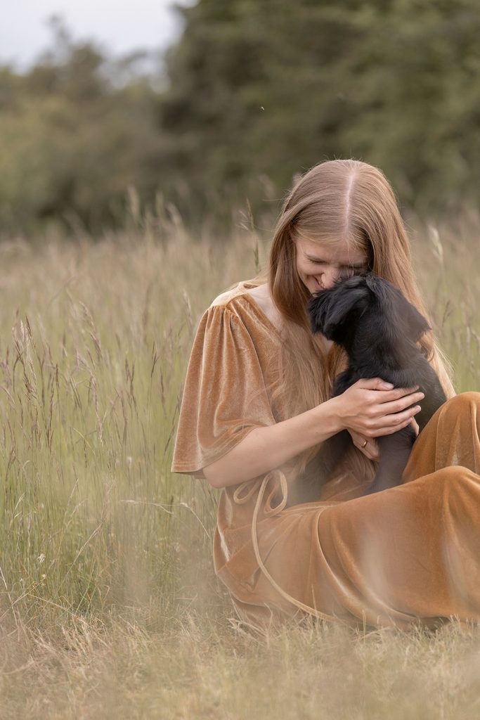 Frau in goldgelben Kleid sitzt mit ihrem Mittelschnauzer Welpen auf einer Wiese im Sommer