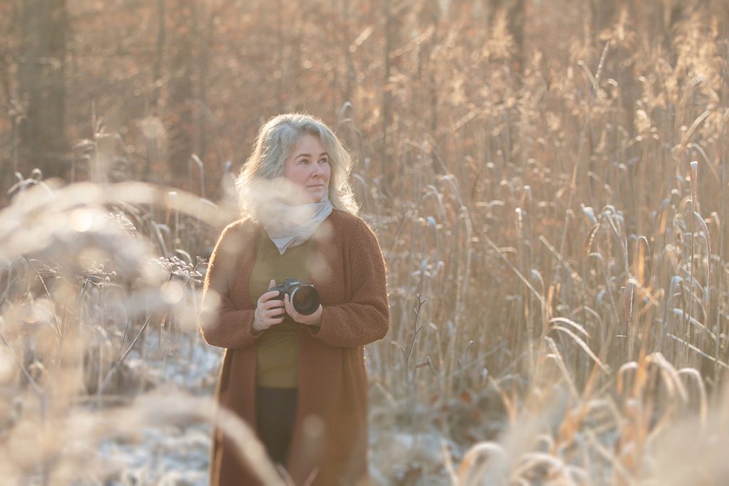Fotografin Sonja mit Kamera in der Hand in einer Schilflandschaft, trägt eine olivfarbene Strickjacke, T-Shirt und schwarze Hose – Wintershooting Stuttgart.