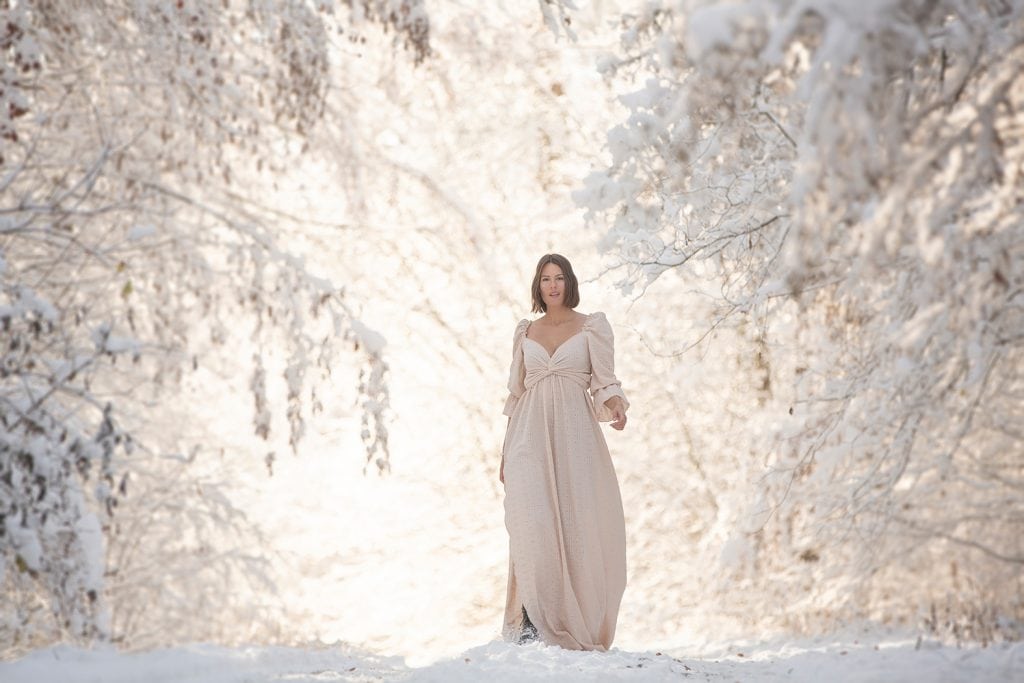 Frau in einem cremefarbenen Shootingkleid läuft durch einen verschneiten Waldweg mit schneebedeckten Ästen bei einem Wintershooting.