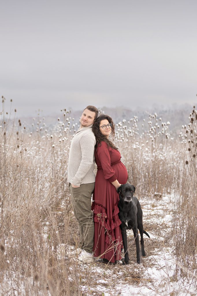 Paar mit schwarzem Hund in einem verschneiten Diestelfeld, Winter Babybauch-Fotoshooting