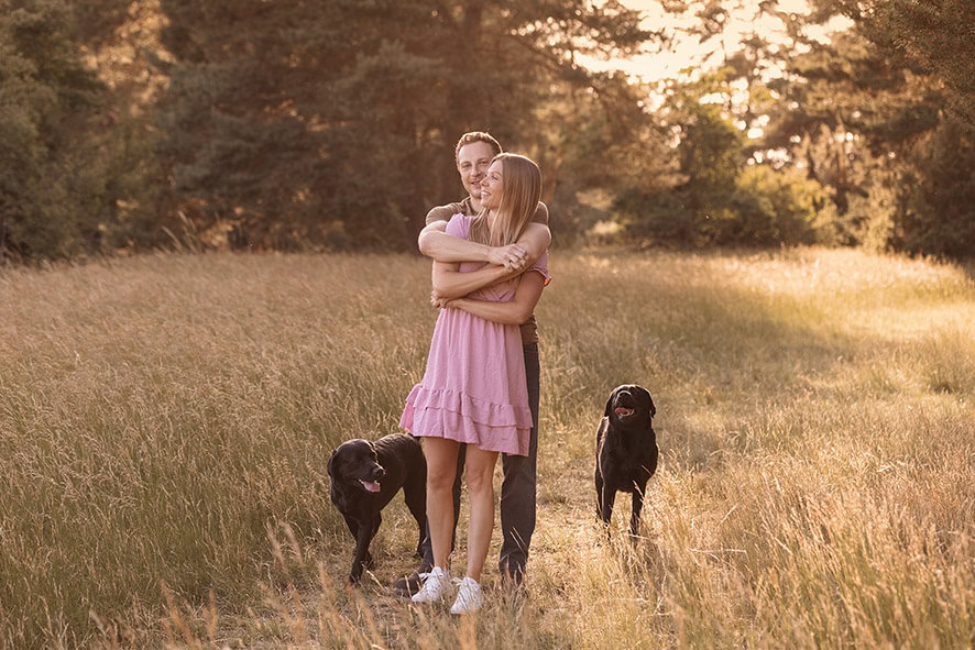 Paar Luft Hand in Hand mit zwei schwarzen Labrador über eine Sommer Wiese im Sonnenuntergang. Stimmungsvolle Aufnahme bei einem Footoshooting mit Sonja Auer Fotografie
